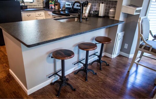 A kitchen with a dark countertop, two stools, and a sink. - Ouachita Weekender 2BR/2BA Condo, Mountain Harbor Resort on Lake Ouachita (Mount Ida)