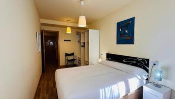 Un dormitorio con cama, mesita de noche y lámpara de pared. Hay una silla en la esquina y una puerta que da a otra habitación.