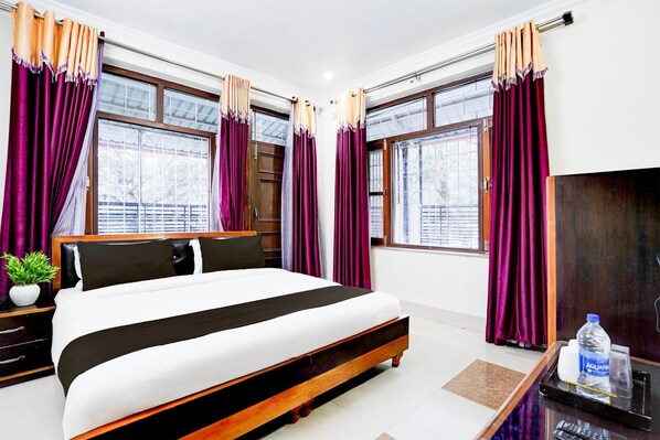 A hotel room with a large bed, purple curtains, a wooden headboard, a nightstand with a plant, and a desk with a water bottle.