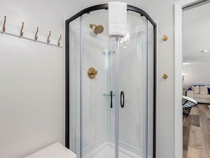A glass-enclosed shower with a towel hanging on the door, a white wall with a towel rack, and a glimpse of a living area with a sofa and a coffee table.