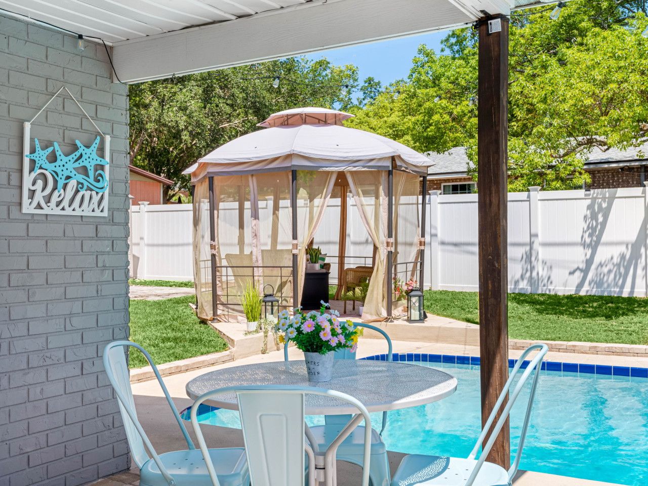 A backyard with a swimming pool, a covered patio, and a table with chairs.