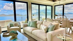Un salon moderne avec un grand canapé, une table basse en verre et une vue sur la mer grâce à de grandes fenêtres.