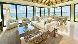 Un vaste salon doté d’un haut plafond, de grandes fenêtres offrant une vue sur la mer, et d’un coin salon confortable avec une table basse et des coussins.