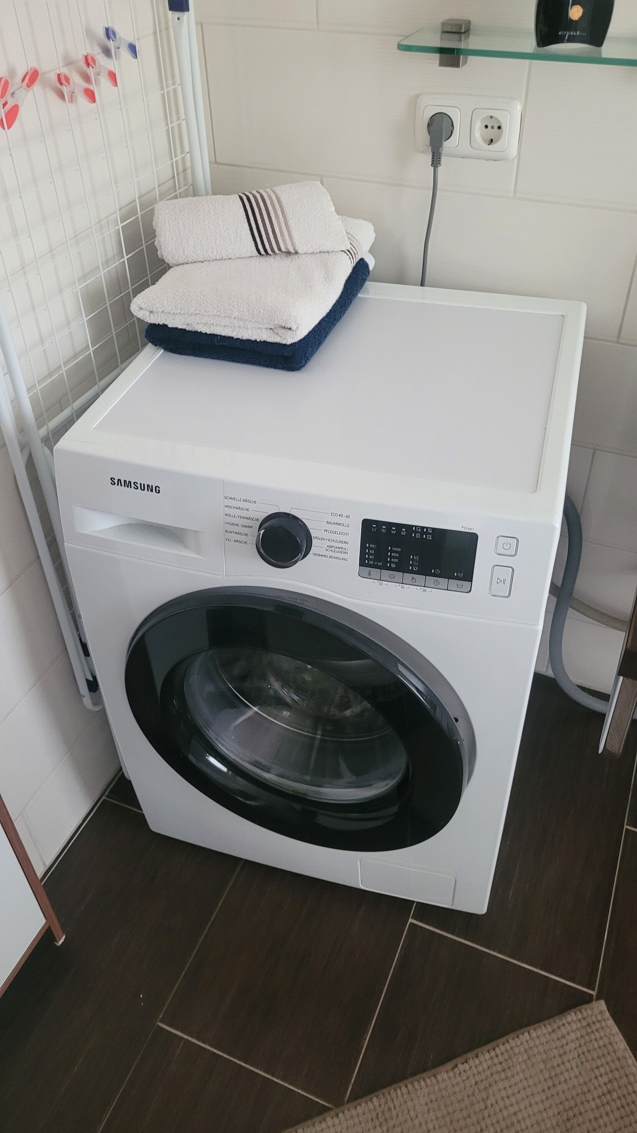 A Samsung front-loading washing machine with a control panel, a towel on top, and a power outlet nearby.