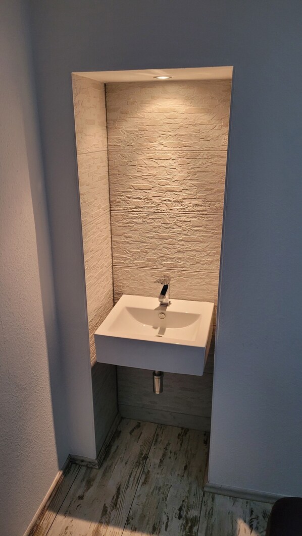 A corner bathroom with a white sink, textured stone wall, and wooden floor. - Beautiful apartment, on the first floor of my own home, with garden access and pool (Zörbig)