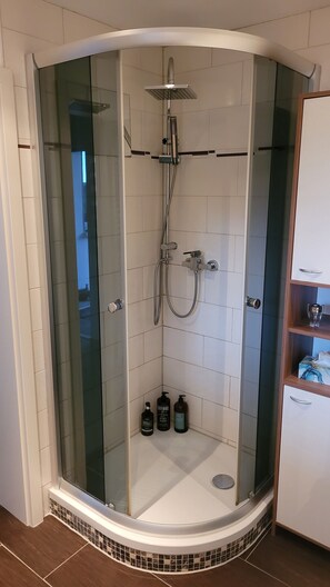 Eine Eckdusche mit Glasabtrennung, eine Handbrause, ein wandmontierter Wasserhahn und eine kleine Ablage mit Toilettenartikeln.