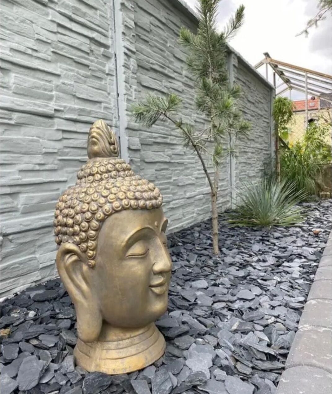 A Buddha statue in a garden setting with a stone wall and a small tree.