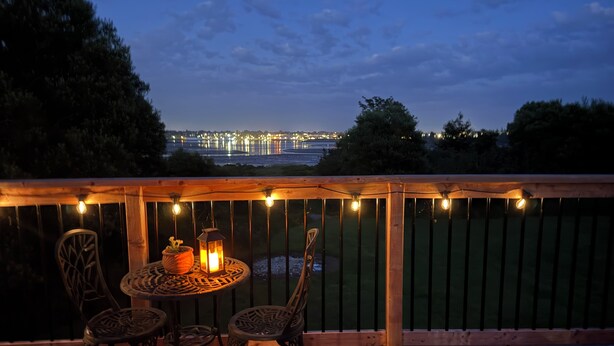 A wooden deck with a table and chairs, string lights, and a view of a body of water with lights in the distance. - Ocean view house (Overton)
