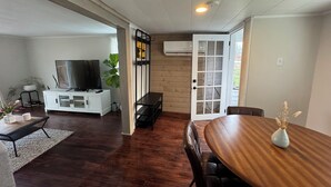 A spacious living area with a wooden dining table, a TV on a white cabinet, and a large window with blinds. - Ocean view house (Overton)