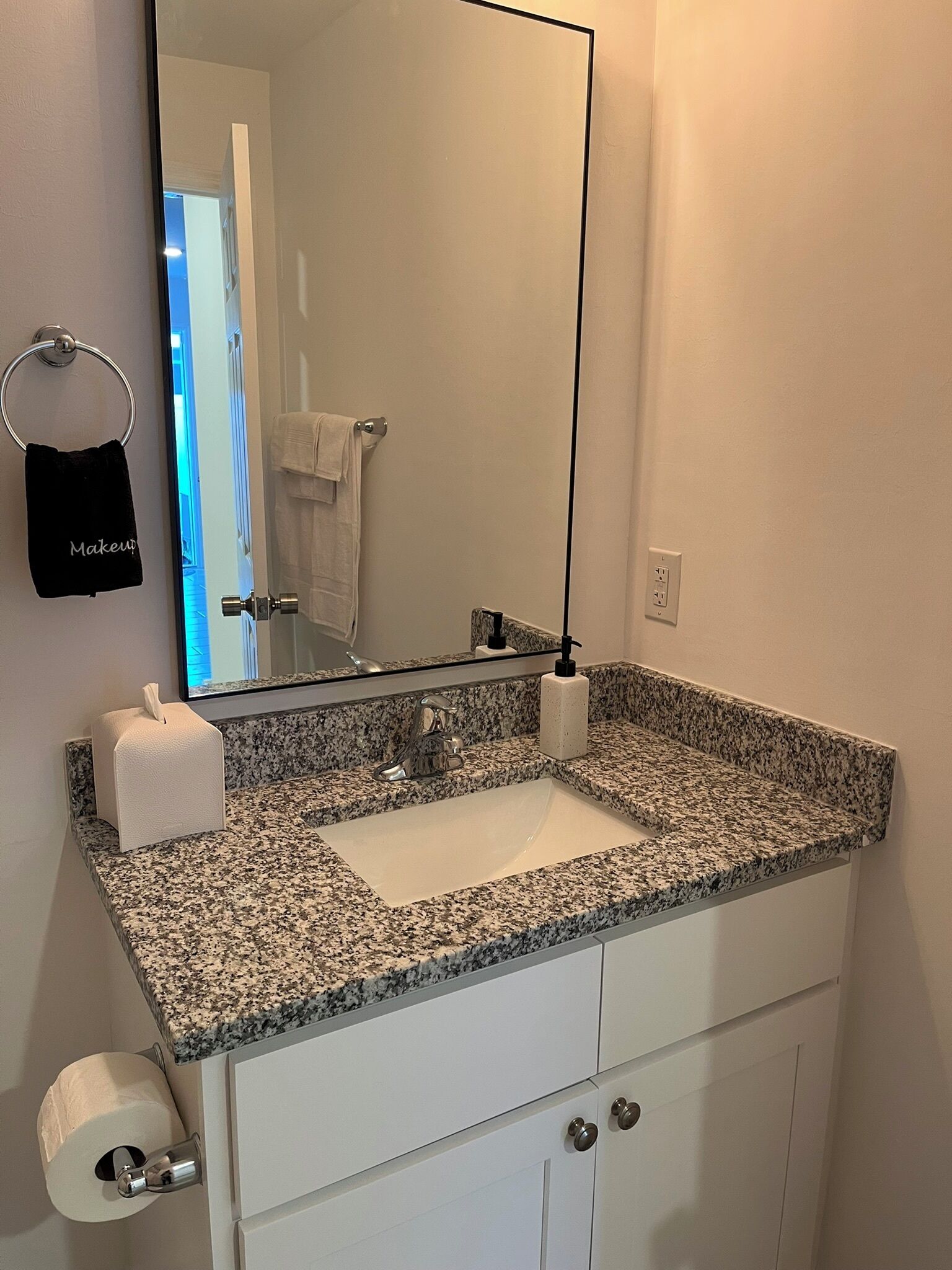 A bathroom with a granite countertop sink, a mirror, a towel rack, a soap dispenser, and a toilet paper holder.