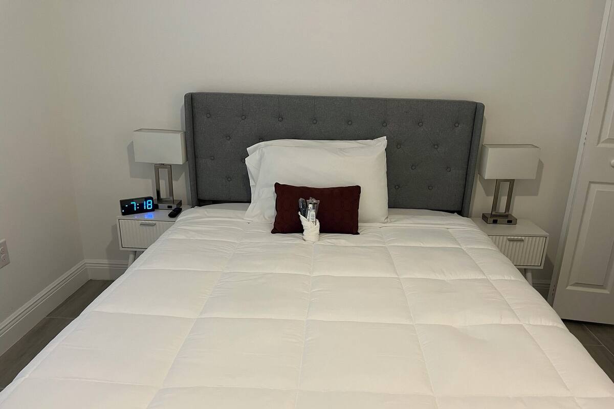 Cama bem arrumada com cabeceira estofada cinza, roupa de cama branca e duas luminárias de cabeceira.