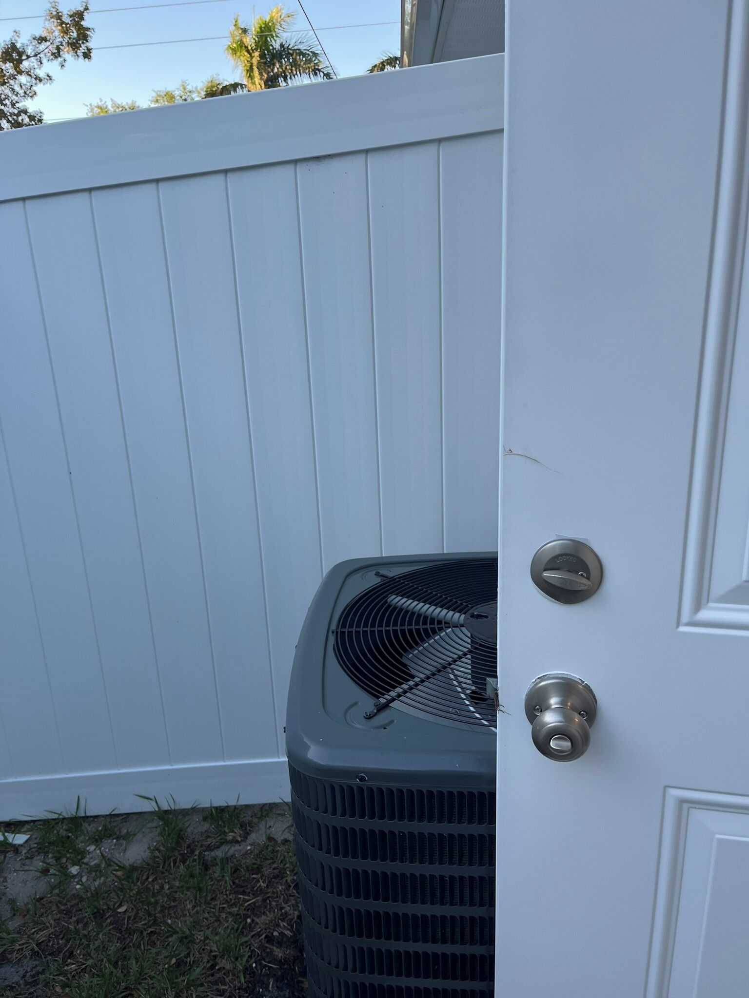 A white door with a metal handle and a keyhole, a black outdoor unit with a grill, and a white fence in the background.