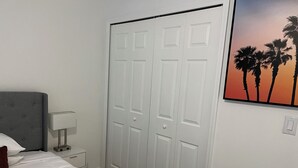A bedroom with a bed, a bedside table with a lamp, a wardrobe, and a sunset painting on the wall. - Cozy Lehigh Extended Stay (Lehigh Acres)