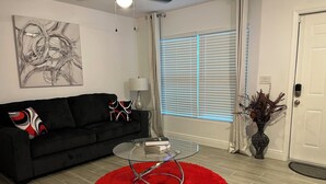 A modern living room with a black sofa, a glass coffee table, a red rug, a painting on the wall, and a door with a keypad lock. - Cozy Lehigh Extended Stay (Lehigh Acres)