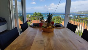 A wooden table with a fruit bowl, red and black cups, and a plate in a balcony with a view of the sea and boats.
