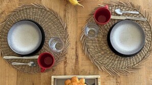 Una mesa de madera con dos individuales tejidos, dos platos, dos tazas rojas, un cuenco de fruta y una bandeja de pasteles.