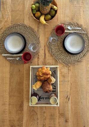 Een houten tafel met twee gevlochten placemats, twee borden, twee rode bekers, een fruitschaal en een bord gebak.'