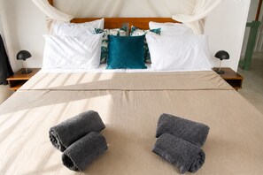 Ein ordentlich bezogenes Bett mit einer beigen Tagesdecke, weißen Kissen und einem Türkis-farbenen Akzentkissen. Es gibt zwei Nachttische mit Lampen und ein Paar aufgerollte Handtücher auf dem Bett.