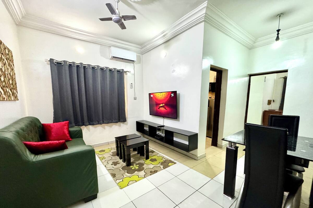A living room with a green sofa, a TV, a ceiling fan, and a dining area with a table and chairs.