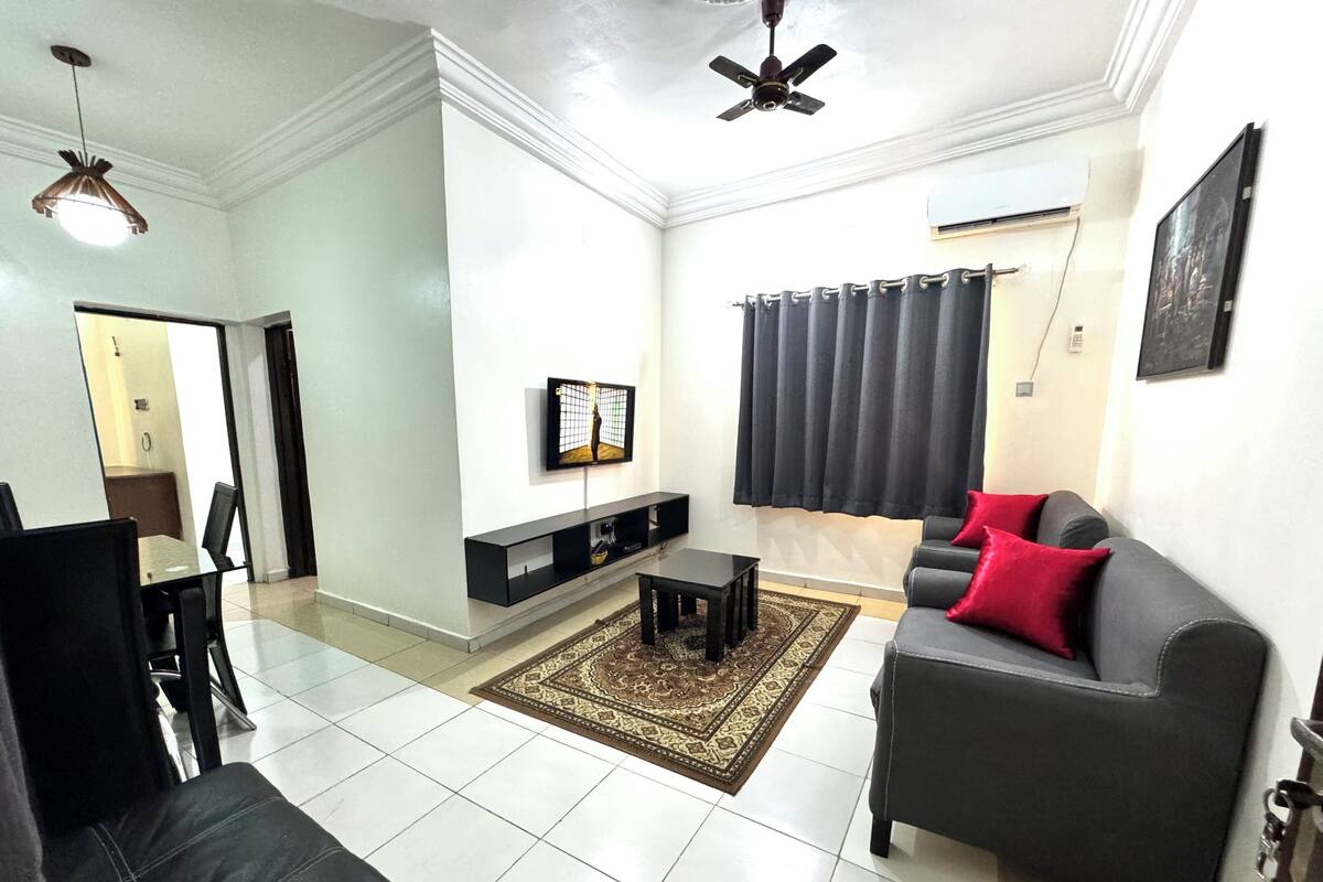A living room with a grey sofa, a TV mounted on the wall, a ceiling fan, and a dining area with a table and chairs.