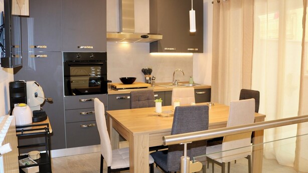 A modern kitchen with a dining area, featuring grey cabinets, a wooden dining table, and a glass railing. - Alumaro House (Castellammare del Golfo)