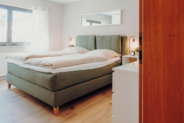 A modern bedroom with a green bed, a wooden nightstand with a lamp, and a large window with curtains. - ApartmentTraveller Saarlouis near city center (Saarlouis)