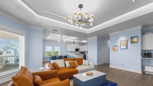 A spacious living room with a modern sofa, a coffee table, and a chandelier. There is a kitchen area in the background with white cabinets and a dining area with a blue rug. - Ocean View, Modern, Elegant Oasis With Private Pool In Gated, Coastal Community! (Eastpoint)
