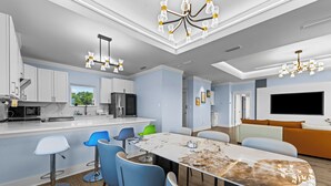 A modern kitchen with white cabinets, a central island, and a dining area with blue chairs and a marble table. - Ocean View, Modern, Elegant Oasis With Private Pool In Gated, Coastal Community! (Eastpoint)