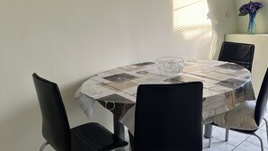 A dining area with a round table covered by a patterned tablecloth, surrounded by four black chairs. There are two framed pictures on the wall, and a vase with purple flowers on a small table to the right.