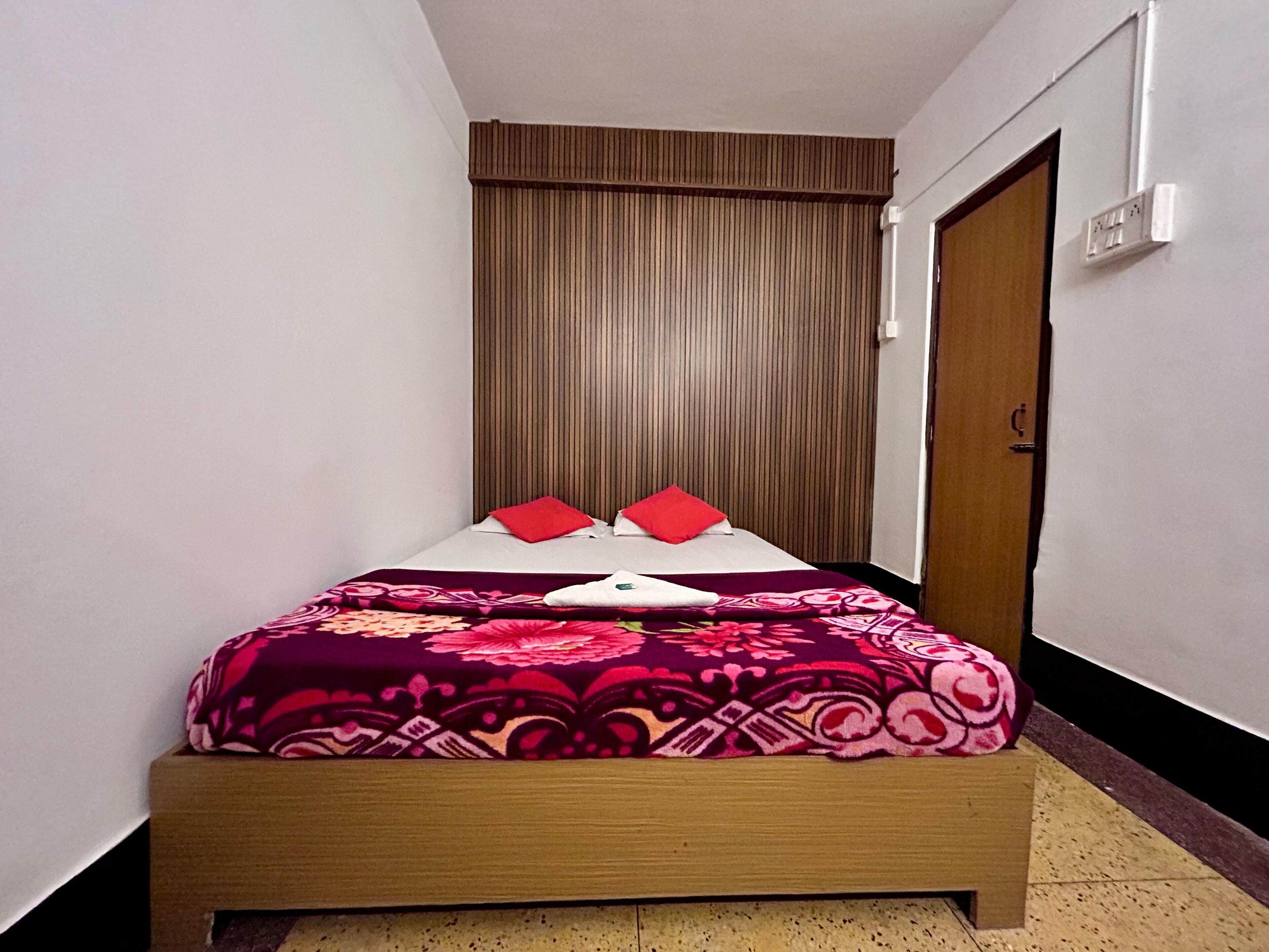 A small, neatly made bed with a patterned bedspread and two red pillows, a wooden headboard, and a door to the right.
