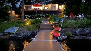 Een modern huis met een houten terras dat uitkomt op een overdekt buitenverblijf, omgeven door een tuin en een vijver.