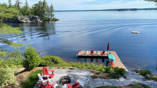 Een terras aan het meer met Adirondack-stoelen, een vuurplaats en een houten steiger die in het water uitloopt.
