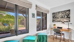 Ein Zimmer mit Schreibtisch, Stuhl und zwei grünen Hocker. Ein Fenster bietet Blick auf grüne Vegetation draußen.