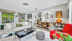 Ein modernes Wohnzimmer mit einer Couch, einem Couchtisch und einem roten Sessel. Ein Bücherregal mit Büchern und Dekorationselementen, sowie ein Fernseher im Hintergrund.