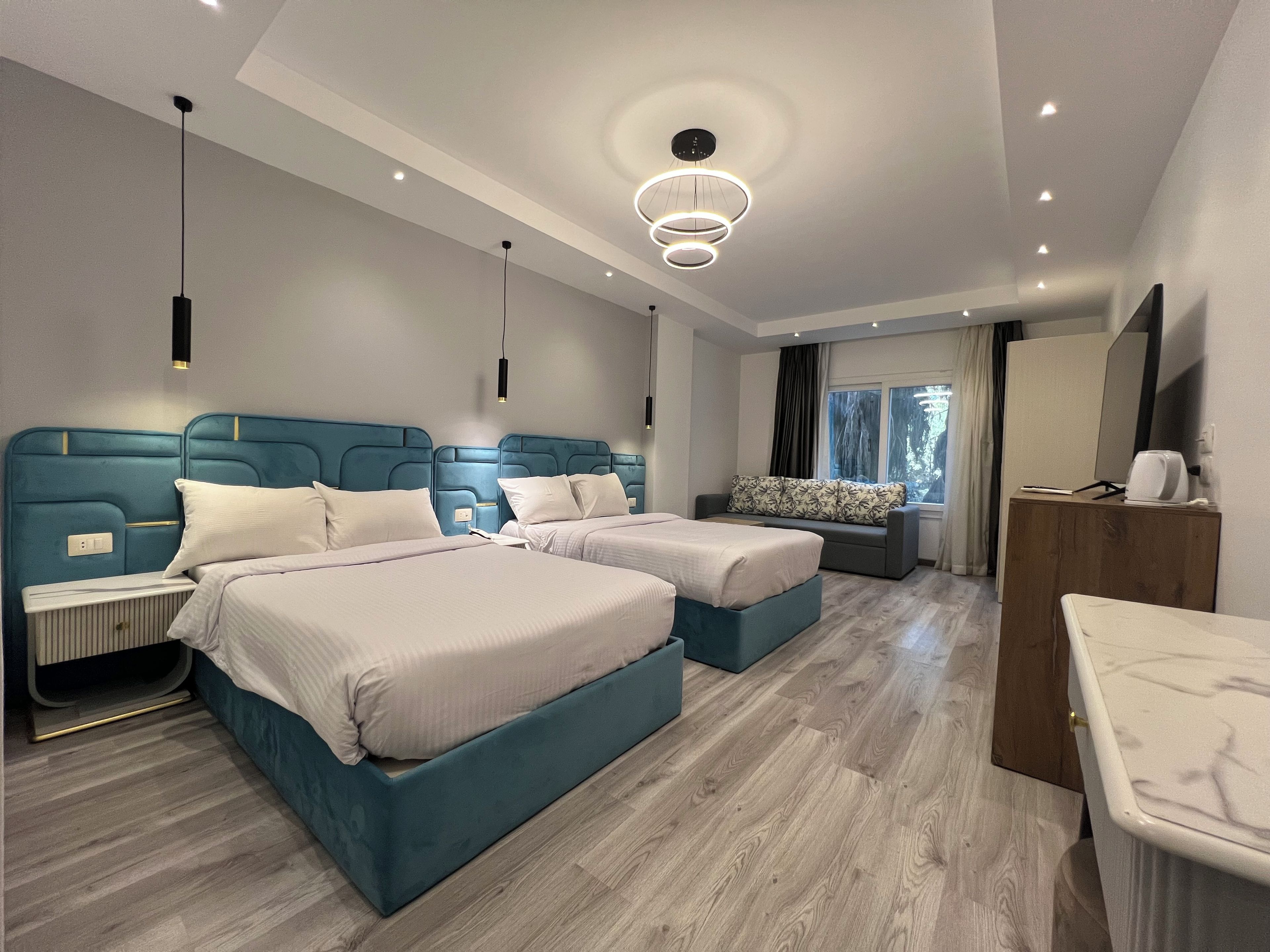 A modern bedroom with two beds, a sofa, a wooden dresser, and a large window.