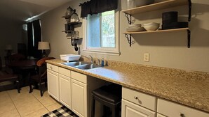 Una cocina con encimera de granito, armarios blancos, alfombra a cuadros blanco y negro, una ventana con cortina negra, y un comedor con mesa y sillas.