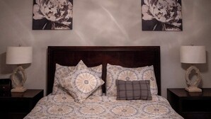 Una cama bien hecha con ropa de cama estampada, dos lámparas fijadas a la pared y dos grandes cuadros florales sobre la cama.