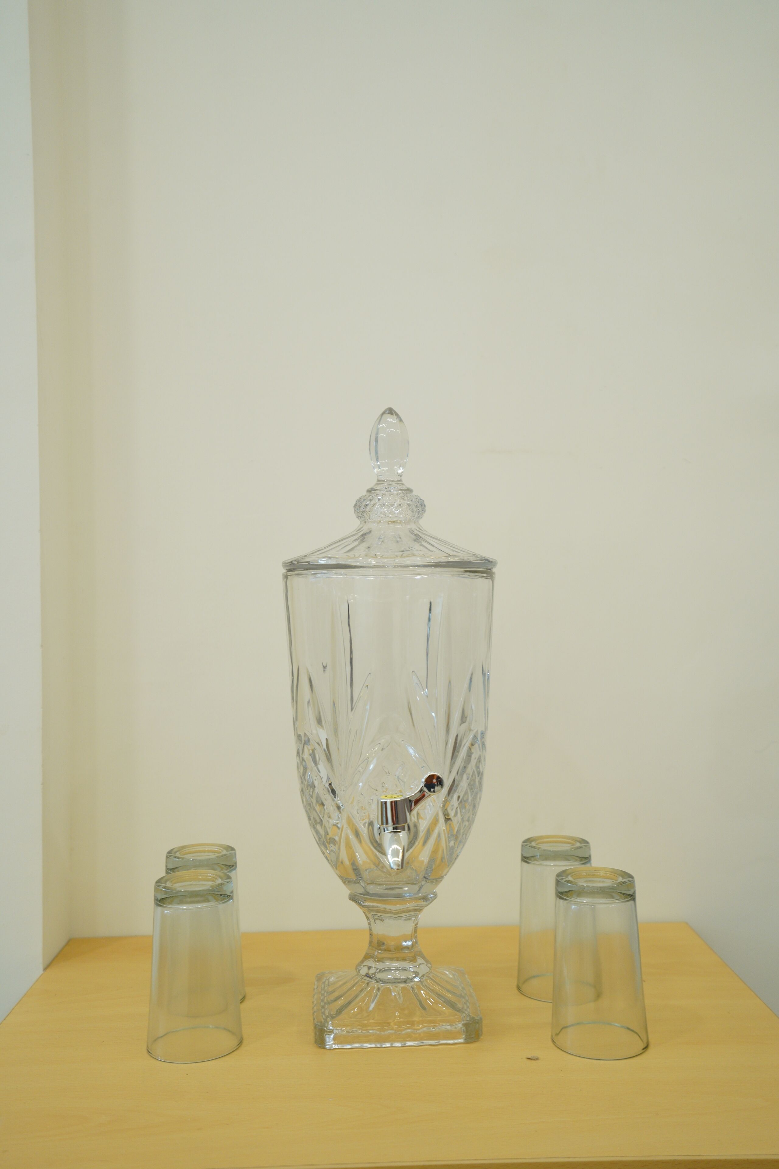 Un bol de ponche de vidrio transparente con tapa y cuatro vasos de vidrio sobre una mesa de madera, frente a una pared lisa.