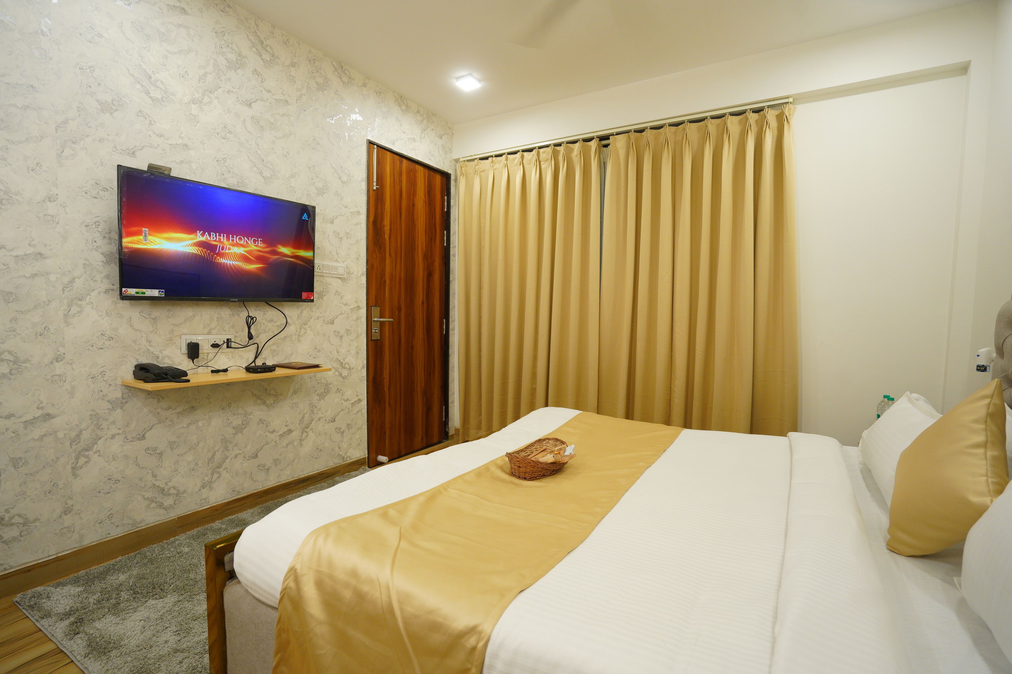 Habitació d'hotel amb un llit, un televisor fixat a la paret, una porta de fusta i cortines beix.