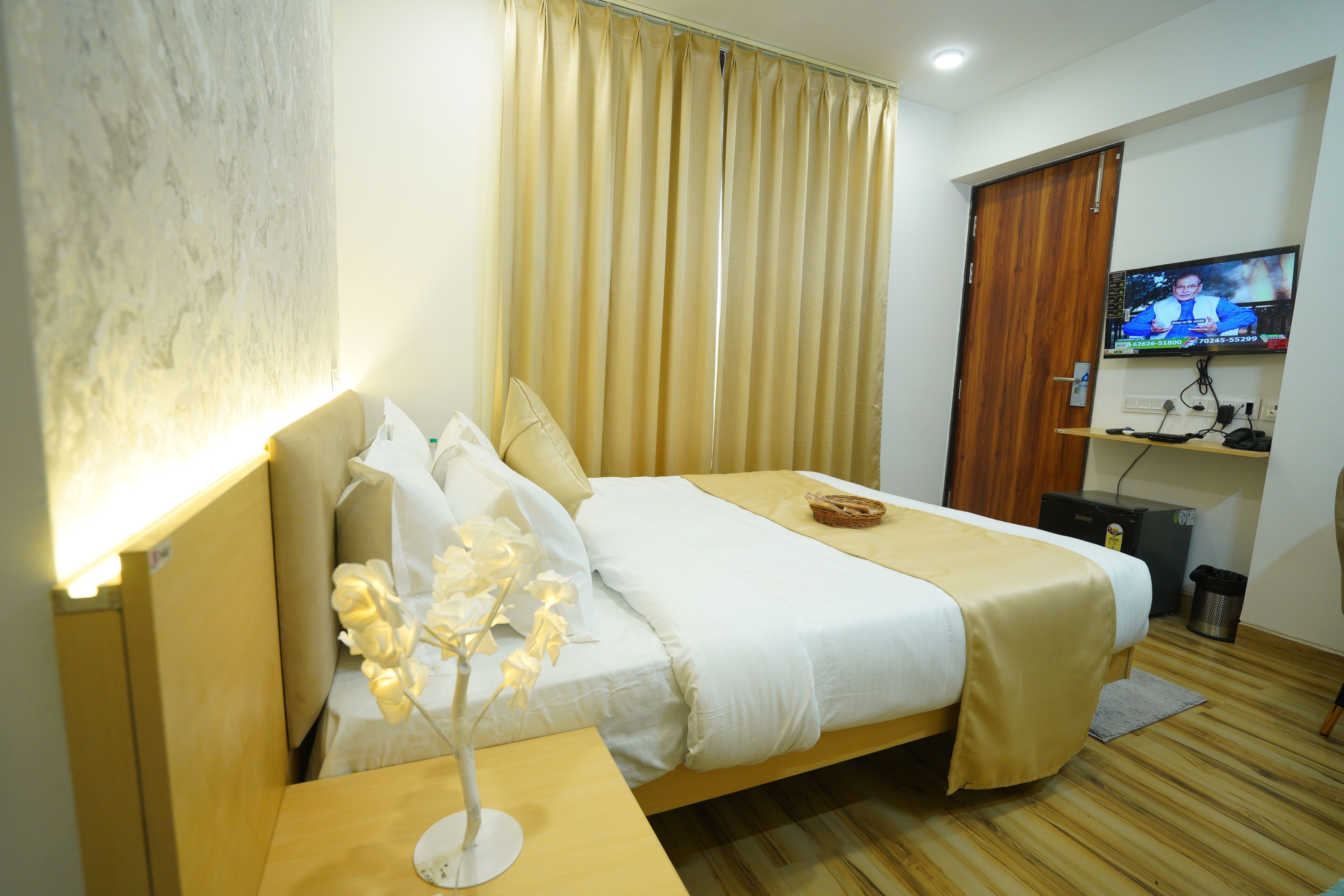 Habitación de hotel con cama, mesita de noche con lámpara, televisor en la pared y piso de madera.