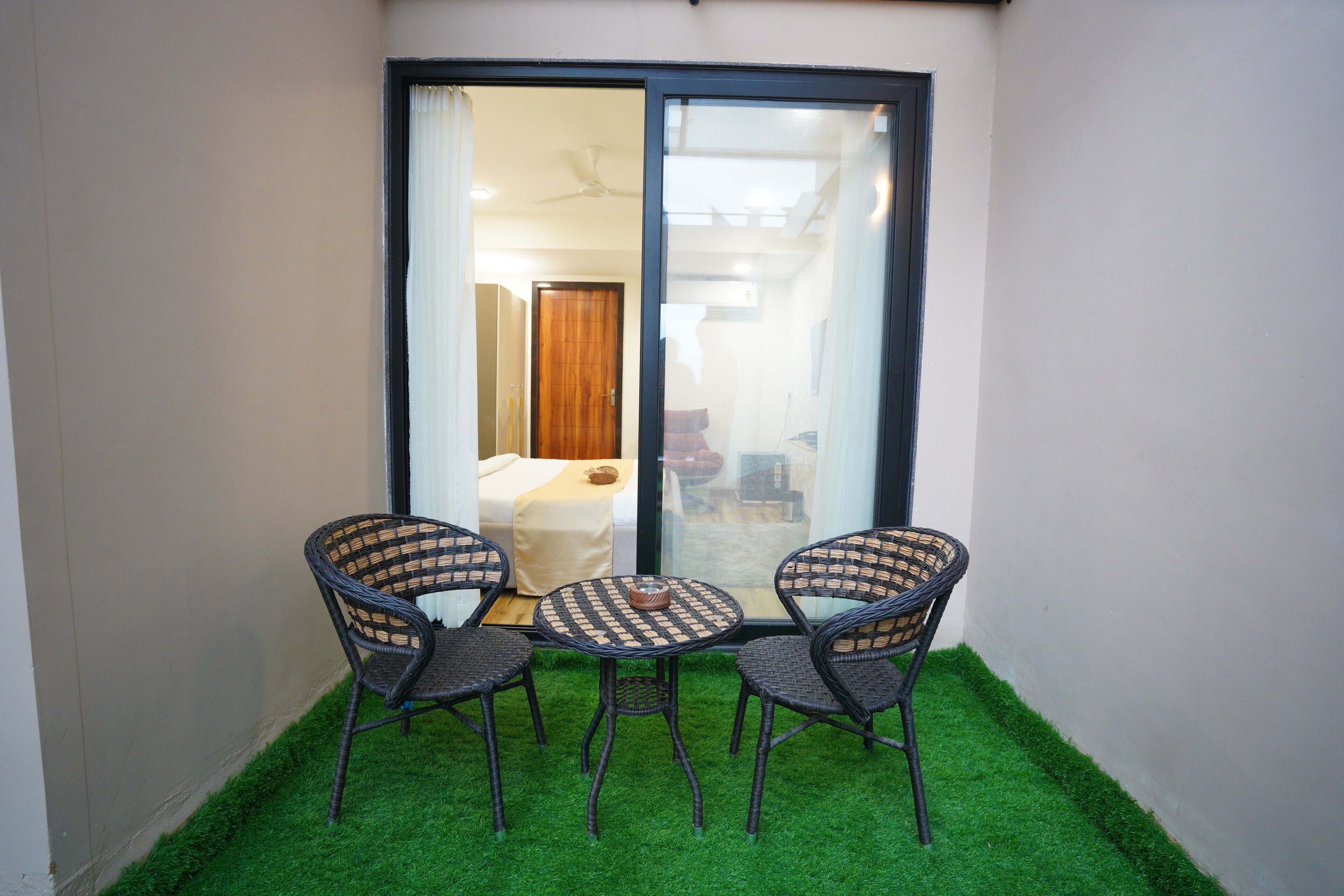 A small balcony with three wicker chairs and a round table, featuring a view into a room with a bed and a wooden door.