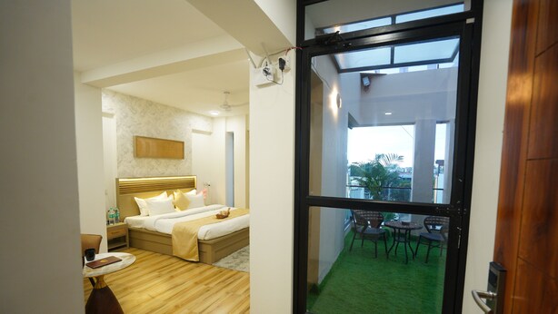 A modern hotel room with a large sliding glass door leading to a balcony with a green lawn and outdoor furniture. - Anugya - A Boutique Hotel (Indore)