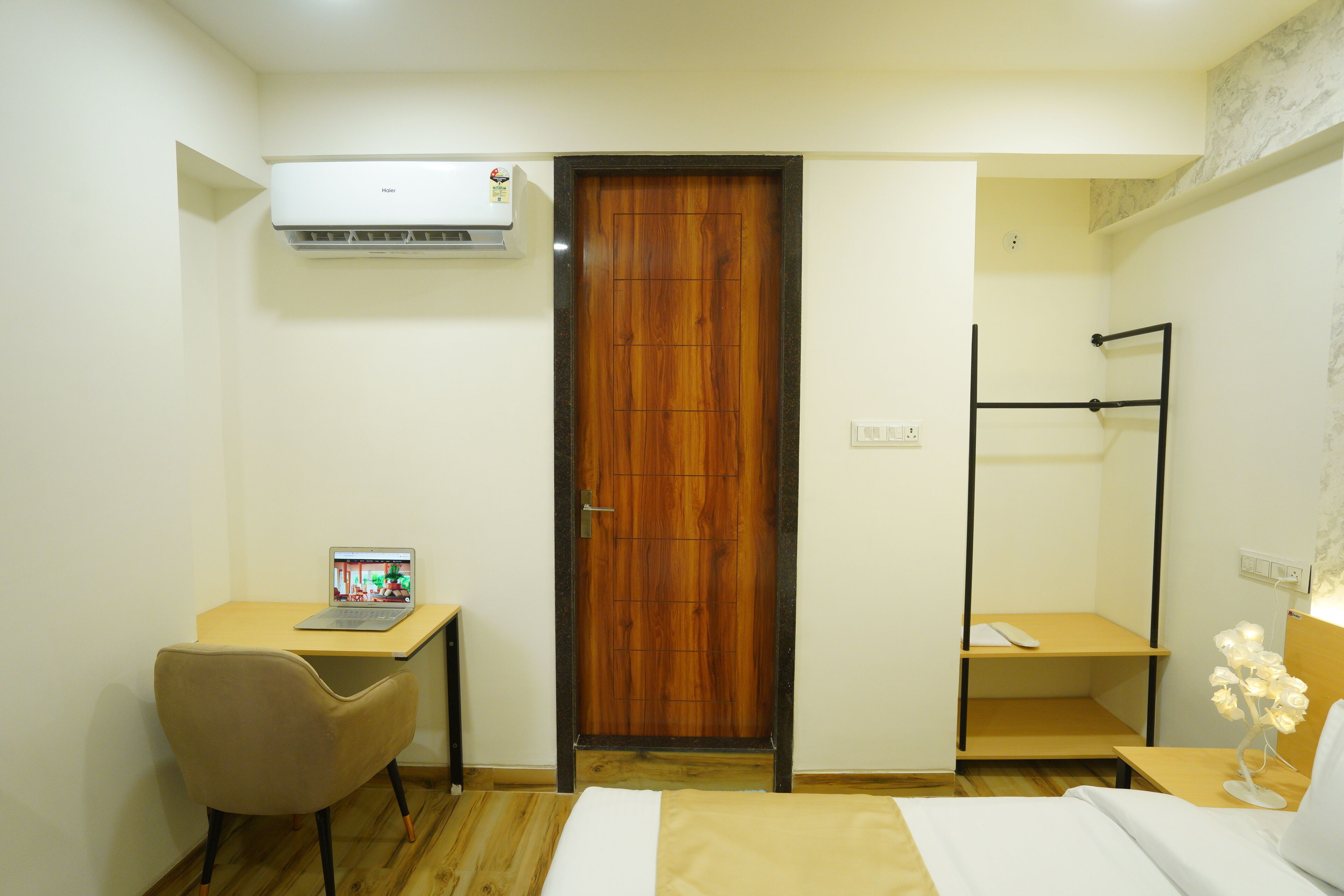 A compact hotel room with a wooden door, a desk with a laptop, a bed with white bedding, and a bedside table with a lamp.