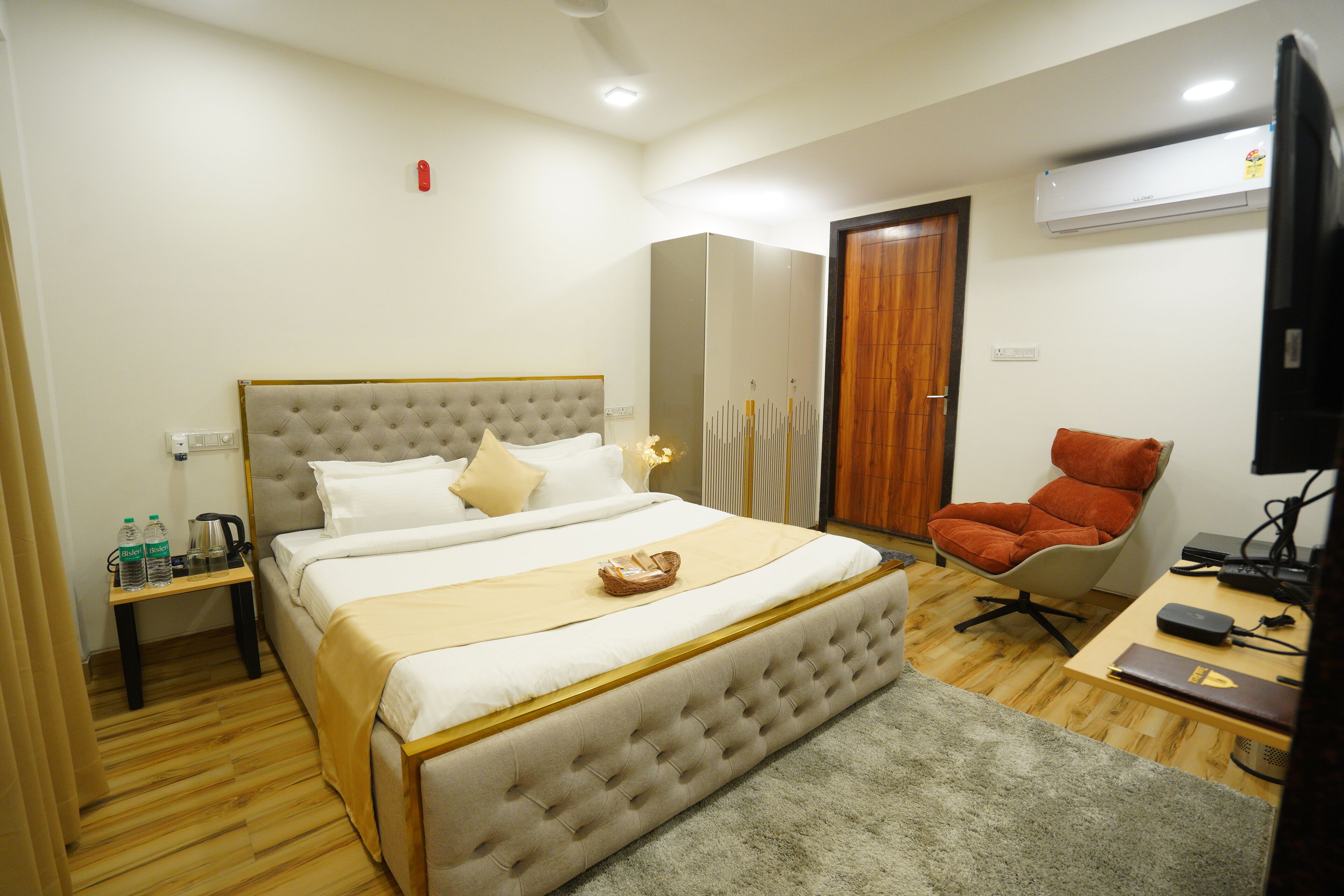 A bedroom with a large bed, a bedside table with a kettle, a chair, a desk with a computer, and an air conditioning unit.