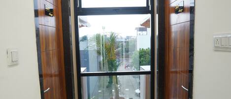 Un pasillo con una puerta de cristal que da a un espacio exterior, puertas de madera a ambos lados, y una planta en maceta cerca de la entrada.