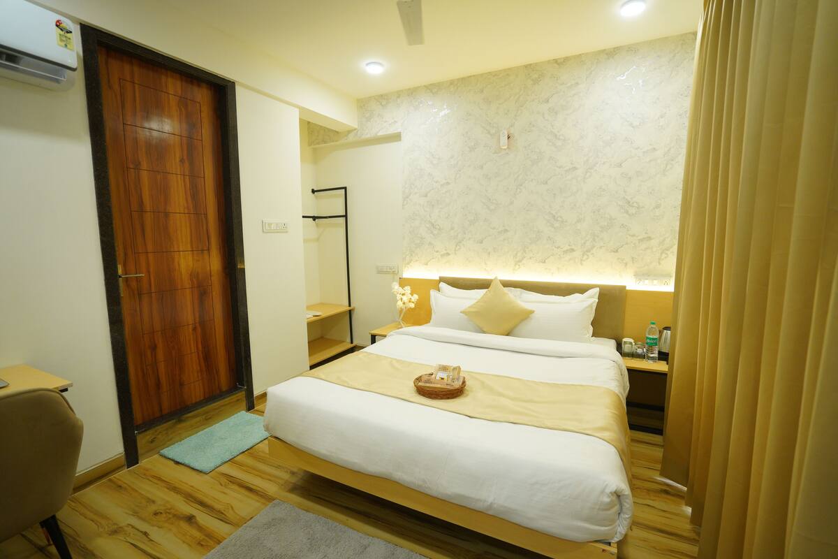 Habitación de hotel con piso de madera, cama con ropa blanca, mesita de noche con lámpara y armario de madera.