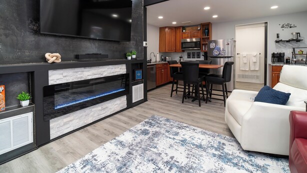 A modern living room with a fireplace, a flat-screen TV, a dining area with a table and chairs, and a kitchen with wooden cabinets and a refrigerator. - The Refined Retreat (Celoron)