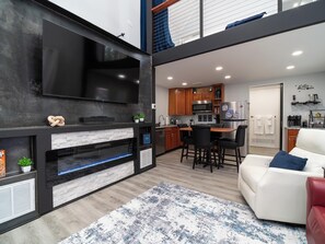 A modern living room with a fireplace, a flat-screen TV, a dining area with a table and chairs, and a kitchen with wooden cabinets and a refrigerator. - The Refined Retreat (Celoron)