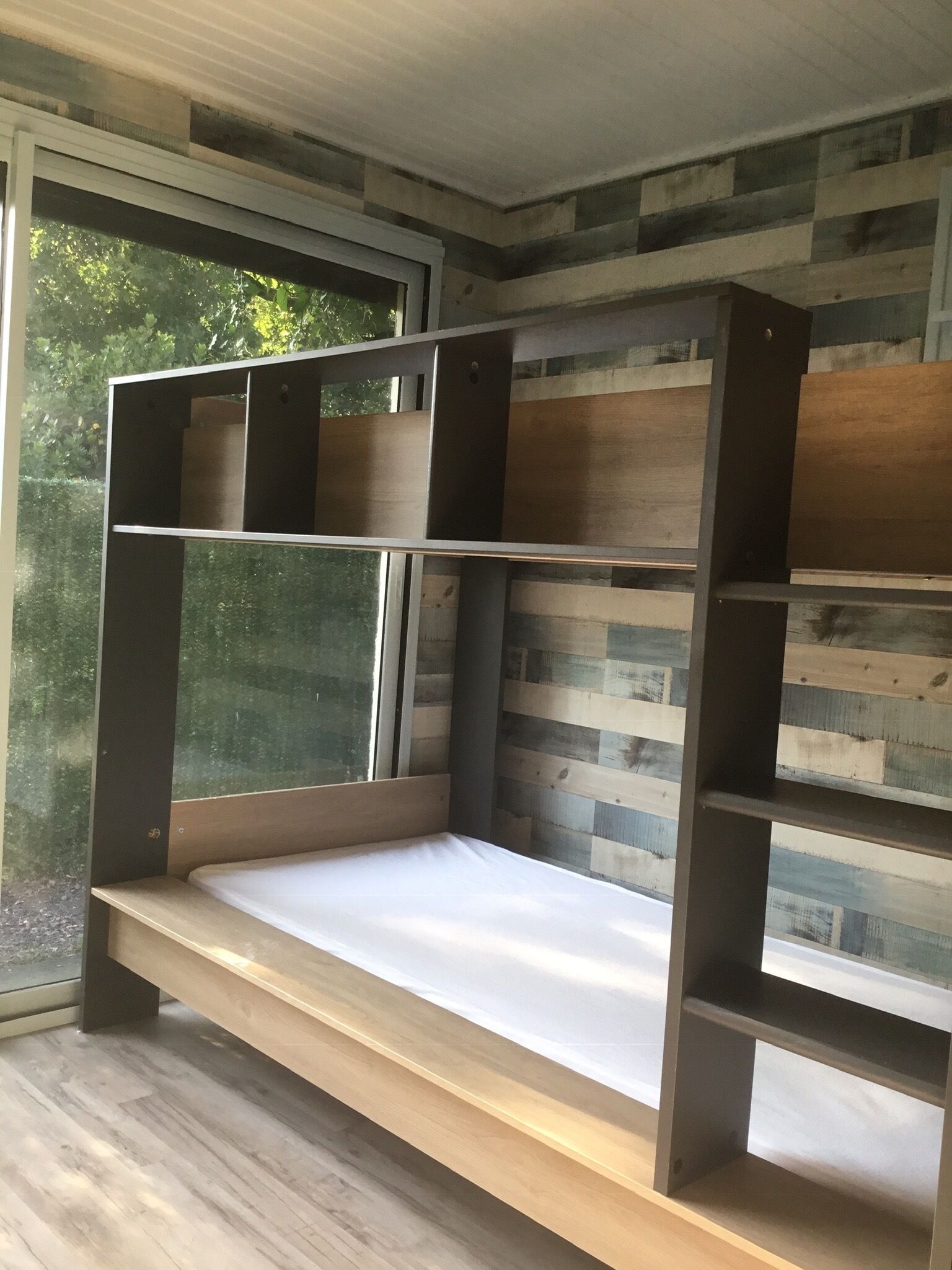 Una cama individual con estructura de madera y colchón, frente a una pared con estampado a rayas.
