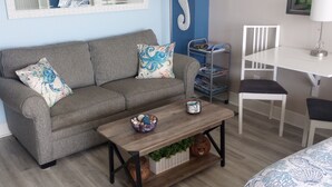 Un canapé gris agrémenté de coussins à motifs floraux, une table basse en bois ornée d’objets décoratifs, un bureau blanc avec une lampe et une chaise blanche.
